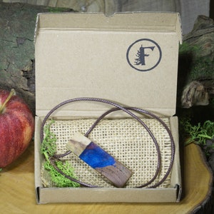 May include: A wooden pendant with blue resin inlay, on a brown cord, in a cardboard box with a brown burlap lining. The box has a logo with a tree and the letter "F".