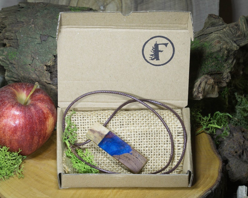 May include: A wooden pendant with blue resin inlay, on a brown cord, in a cardboard box with a brown burlap lining. The box has a logo with a tree and the letter "F".