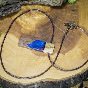 May include: A brown leather cord necklace with a wooden pendant featuring a blue and purple resin inlay. The pendant is rectangular and has a natural wood grain texture.