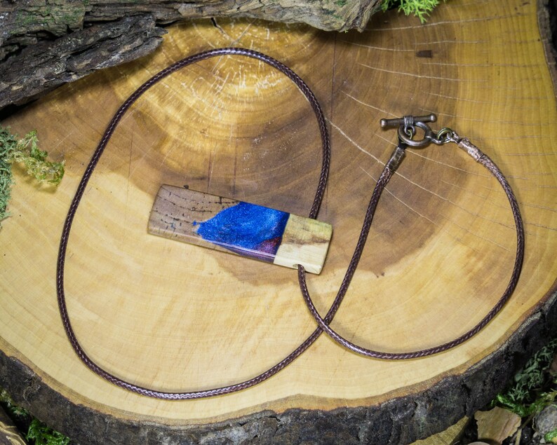 May include: A brown leather cord necklace with a wooden pendant featuring a blue and purple resin inlay. The pendant is rectangular and has a natural wood grain texture.