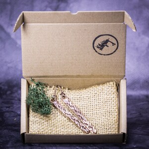 May include: A pair of copper earrings in a cardboard box with a burlap pouch. The earrings are long and have a braided design.