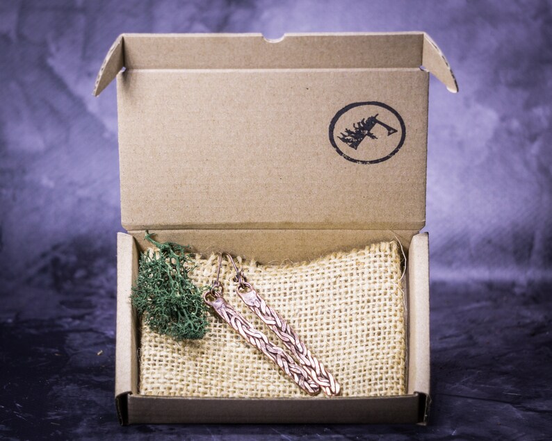 May include: A pair of copper earrings in a cardboard box with a burlap pouch. The earrings are long and have a braided design.