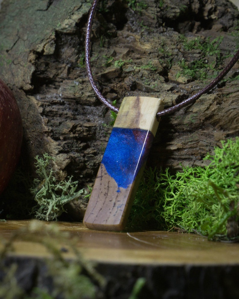 May include: A wooden pendant with a blue and purple resin inlay. The pendant is attached to a brown cord.