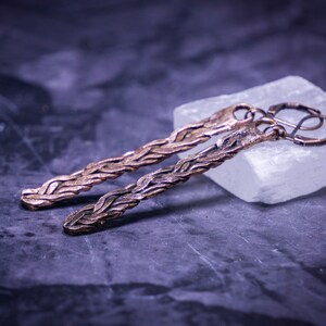 May include: A pair of long, braided copper earrings with a hook closure. The earrings are displayed on a white surface.
