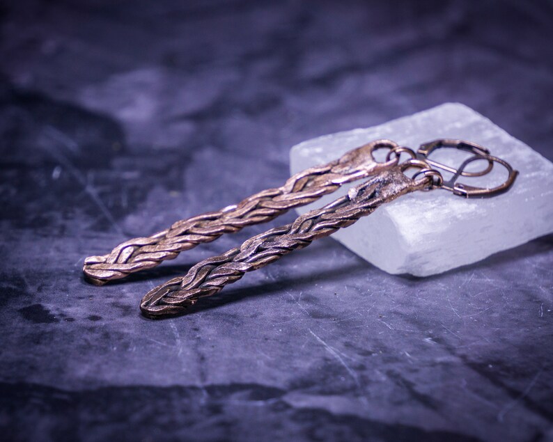 May include: A pair of long, braided copper earrings with a hook closure. The earrings are displayed on a white surface.