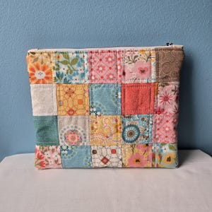 May include: A patchwork zippered pouch with a variety of floral and geometric patterns in shades of pink, yellow, blue, and green. The pouch is rectangular and has a white zipper and a white lining.