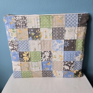 May include: A patchwork zippered pouch with a variety of floral and geometric patterns in shades of blue, green, brown, and cream. The bag is made of quilted fabric and has a white zipper.