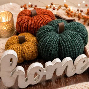 May include: Three knitted pumpkins in orange, mustard yellow, and dark green, each featuring a brown leather-like stem. A white wooden cutout sign with the word home is placed in front of the pumpkins on a dark brown tray.