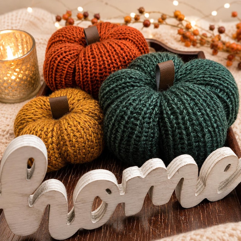 May include: Three knitted pumpkins in orange, mustard yellow, and dark green, each featuring a brown leather-like stem. A white wooden cutout sign with the word home is placed in front of the pumpkins on a dark brown tray.