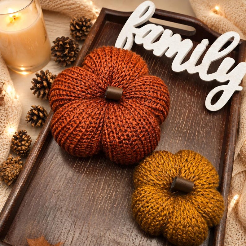 May include: Two knitted pumpkins in rust orange and mustard yellow, each featuring a small brown stem. The pumpkins are placed on a dark brown wooden tray alongside a white wooden cutout sign with the word family in cursive script.