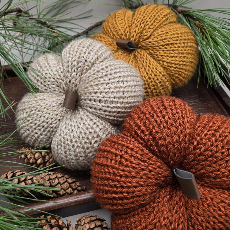 May include: Three knitted pumpkins in beige, orange, and yellow. The pumpkins are all the same size and have a similar texture. They are all sitting on a wooden surface.
