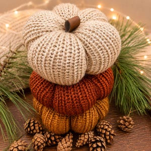 May include: A stack of three knitted fabric pumpkins in beige, burnt orange, and mustard yellow. Each pumpkin features a ribbed texture and a small brown leather-like stem at the top center. The pumpkins are arranged vertically as a decorative set.