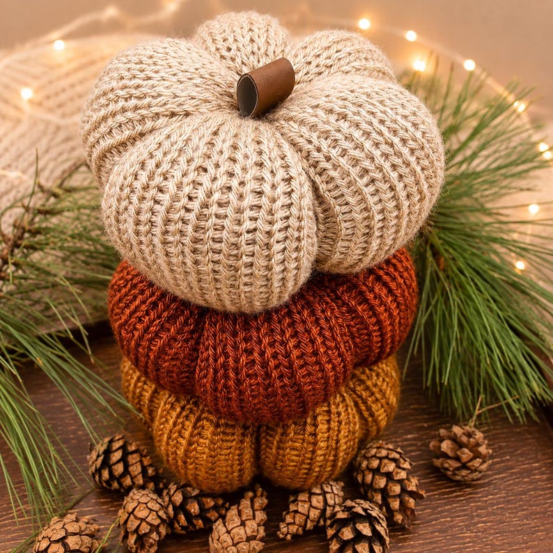 May include: A stack of three knitted fabric pumpkins in beige, burnt orange, and mustard yellow. Each pumpkin features a ribbed texture and a small brown leather-like stem at the top center. The pumpkins are arranged vertically as a decorative set.