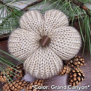 May include: A knitted pumpkin in a light tan color labeled Grand Canyon, featuring a ribbed texture and a smooth, dark brown cylindrical stem at the center. The pumpkin is crafted with a visible knit pattern and has a rounded, segmented shape.