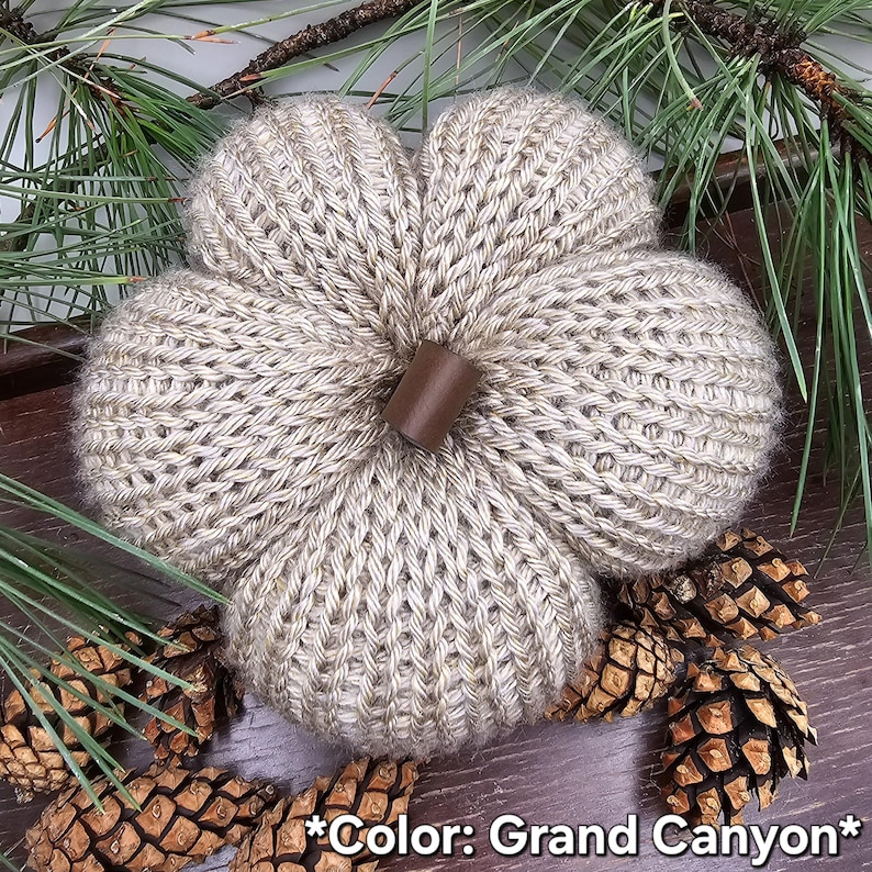 May include: A knitted pumpkin in a light tan color labeled Grand Canyon, featuring a ribbed texture and a smooth, dark brown cylindrical stem at the center. The pumpkin is crafted with a visible knit pattern and has a rounded, segmented shape.