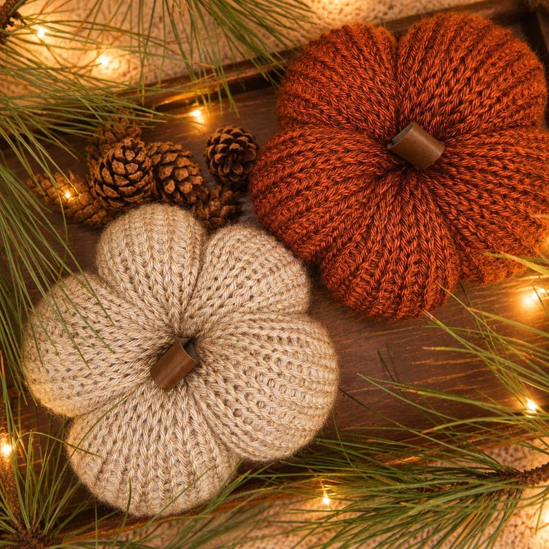 May include: Two knitted pumpkins, one in a deep rust orange and one in a light beige, each featuring a smooth brown stem. The pumpkins are arranged alongside several small brown pinecones and green pine needles on a dark wooden surface.