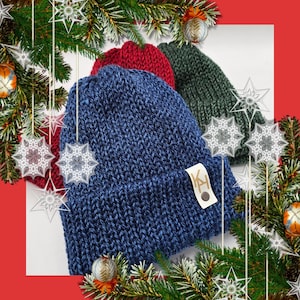 May include: Three knitted beanies in blue, red, and green are displayed. The blue beanie is in the foreground, with a small tag attached. The beanies are surrounded by Christmas decorations, including pine branches, ornaments, and snowflake accents.