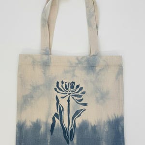 May include: A canvas tote bag with a blue and white tie-dye effect. The bag features a dark blue floral design in the center. The handles are the same color as the bag.