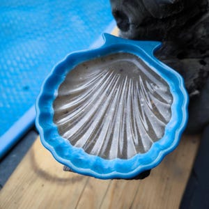 Könnte beinhalten: Eine blaue, muschelförmige Schale gefüllt mit Sand. Die Schale hat einen gewellten Rand und ein strukturiertes Inneres, das die natürlichen Linien einer Muschel nachahmt. Die Schale liegt auf einer Holzoberfläche.