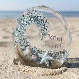 Könnte beinhalten: Ein klarer, facettierter Glasornament mit Strandmotiv. Im Inneren bilden silberne Glitzer ein Herz, mit Muscheln und einem Seestern an der Basis. Der Text "Meer Zeit" ist sichtbar. Der Ornament liegt auf Sand.