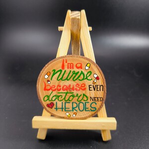 Könnte beinhalten: Ein rundes Holzschild mit dem Text "I'm a nurse because even doctors need heroes", der in bunten Buchstaben bemalt ist. Das Schild ist mit kleinen roten und gelben Pillen und einem roten Herzen verziert. Das Schild wird auf einem kleinen Holzständer präsentiert.
