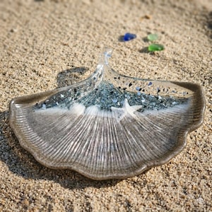 Könnte beinhalten: Eine dekorative, blattförmige Schale mit Küstenmotiv. Die Schale hat eine sandbraune Basis mit einem strukturierten, fächerartigen Muster. Der obere Teil hat ein blau-weißes Design mit eingebettetem Glitzer und einem Seestern. Die Schale liegt auf Sand.
