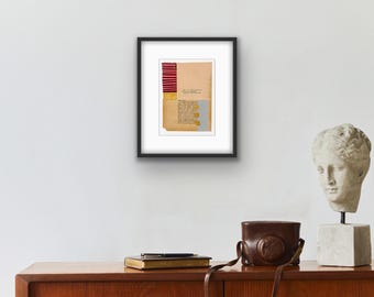 Puede incluir: Obra de arte enmarcada con un collage de papel, que incorpora elementos rojos, dorados y azules. La pieza cuelga de una pared blanca, complementada con un busto y una funda de cámara de cuero sobre una superficie de madera.
