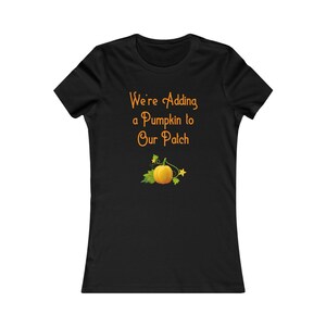 May include: Black t-shirt with the text "We're Adding a Pumpkin to Our Patch" in orange script. Below the text is a graphic of a pumpkin with green leaves and a star. The shirt is a fitted style.