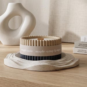 May include: A cylindrical candle with a ribbed exterior, featuring a two-tone design with a tan top and dark blue bottom. A paper label wraps around the centre with the text maple noir | maple syrup & sugar. It sits on a textured, wavy white base.