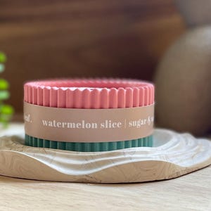Puede incluir: Una vela rosa y verde con el texto "watermelon slice | sugar &" en una bandeja decorativa marmolada. La vela tiene un dise&ntilde;o acanalado y est&aacute; rodeada por una banda marr&oacute;n claro.