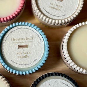 Puede incluir: Primer plano de varias velas de cera de soja en recipientes decorativos. Las velas tienen forma circular con un borde corrugado en varios colores, incluyendo rosa, azul y negro. Cada vela tiene una etiqueta de papel con texto.