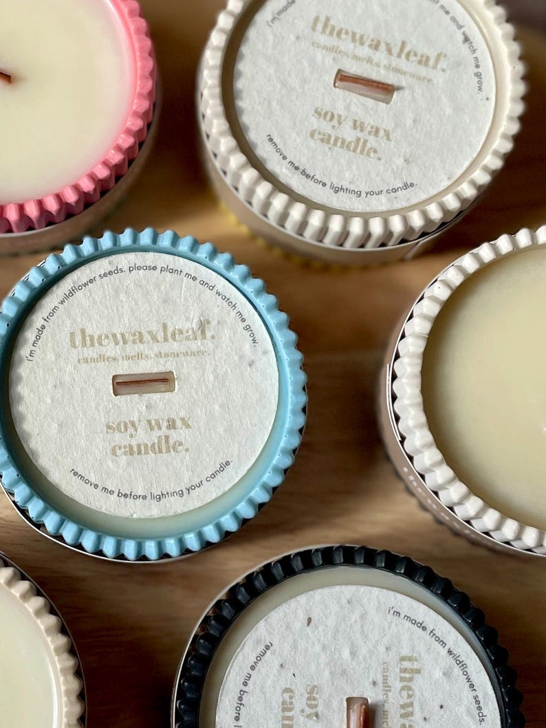 Puede incluir: Primer plano de varias velas de cera de soja en recipientes decorativos. Las velas tienen forma circular con un borde corrugado en varios colores, incluyendo rosa, azul y negro. Cada vela tiene una etiqueta de papel con texto.