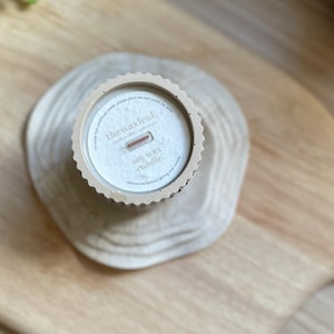 May include: A soy wax candle in a beige container with a scalloped edge, resting on a wooden slice. The candle has a wooden wick and the text "thewaxleaf" is visible. The candle is on a wooden surface.