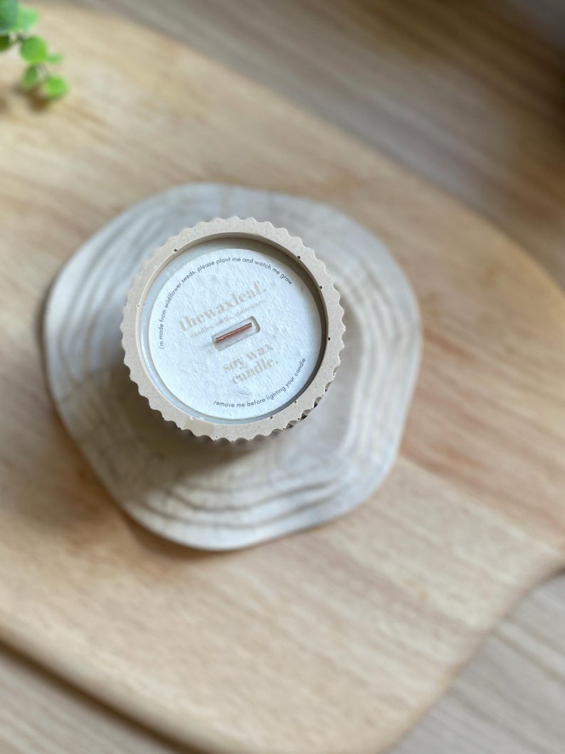 May include: A soy wax candle in a beige container with a scalloped edge, resting on a wooden slice. The candle has a wooden wick and the text "thewaxleaf" is visible. The candle is on a wooden surface.
