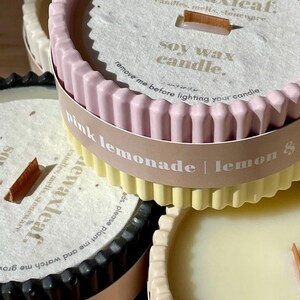 May include: Close-up of several soy wax candles in decorative containers. The candles have a fluted edge and are in various colors, including pink, yellow, and black. Each candle has a wooden wick and a label that reads "Pink Lemonade | Lemon".