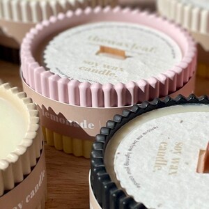 Puede incluir: Primer plano de varias velas de cera de soja en recipientes decorativos. Las velas tienen forma circular con un borde acanalado. Los recipientes son de varios colores, incluyendo rosa, negro y crema. El texto "the wax leaf" y "soy wax candle" es visible.