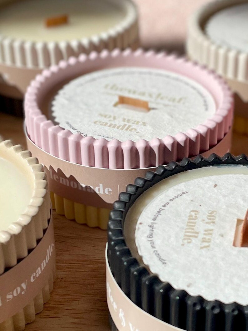 Puede incluir: Primer plano de varias velas de cera de soja en recipientes decorativos. Las velas tienen forma circular con un borde acanalado. Los recipientes son de varios colores, incluyendo rosa, negro y crema. El texto "the wax leaf" y "soy wax candle" es visible.
