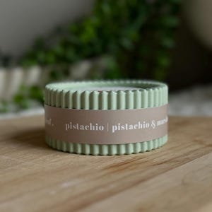 May include: A pale green, circular candle with a ridged design. A brown band encircles the candle, displaying the words "pistachio | pistachio & marsh". The candle is placed on a wooden board.