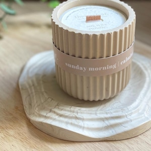 May include: A beige soy wax candle in a ribbed concrete container with a wooden wick, resting on a light-colored, marbled tray. The candle is labeled with the words "sunday morning | calming".