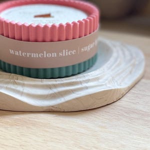 Puede incluir: Una vela con aroma a sand&iacute;a en un recipiente rosa y verde azulado con bordes acanalados, sobre una bandeja blanca veteada. La vela tiene una mecha de madera y cera rosa claro. La etiqueta dice "watermelon slice".