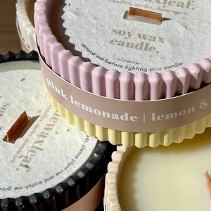 Puede incluir: Primer plano de varias velas de cera de soja en recipientes decorativos. Las velas tienen un borde acanalado y son de varios colores, incluyendo rosa, amarillo y negro. Cada vela tiene una mecha de madera y el texto "pink lemonade | lemon" es visible.