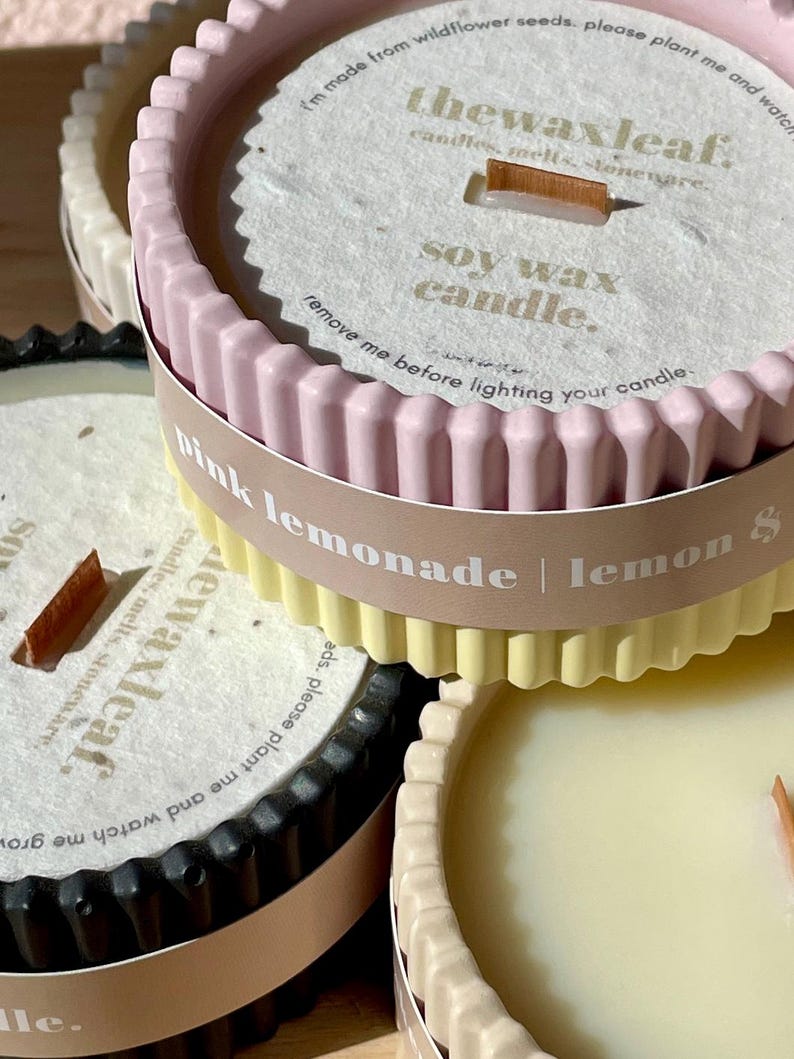 Puede incluir: Primer plano de varias velas de cera de soja en recipientes decorativos. Las velas tienen un borde acanalado y son de varios colores, incluyendo rosa, amarillo y negro. Cada vela tiene una mecha de madera y el texto "pink lemonade | lemon" es visible.