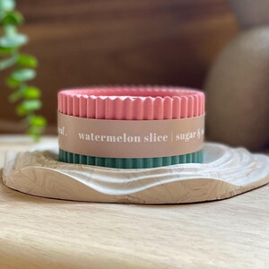 Puede incluir: Una vela rosa y verde con el texto "watermelon slice | sugar &" en una banda marr&oacute;n claro. La vela se encuentra sobre una bandeja decorativa, veteada, blanquecina. La bandeja est&aacute; sobre una superficie de madera.