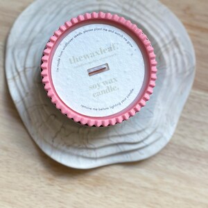 Puede incluir: Una vela de cera de soja en un recipiente rosa y negro, sobre una superficie de madera clara. La etiqueta de la vela dice "thewaxleaf" e incluye el texto "I'm made from wildflower seeds, please plant me and watch me grow."