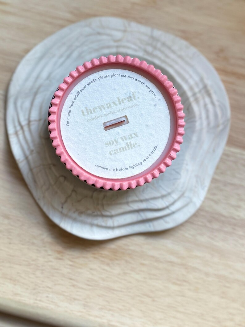 Puede incluir: Una vela de cera de soja en un recipiente rosa y negro, sobre una superficie de madera clara. La etiqueta de la vela dice "thewaxleaf" e incluye el texto "I'm made from wildflower seeds, please plant me and watch me grow."