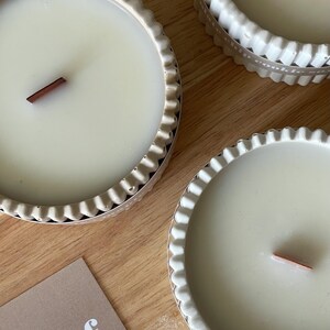 Puede incluir: Primer plano de tres velas de color crema en recipientes decorativos con bordes festoneados. Cada vela tiene una mecha de madera. Las velas est&aacute;n sobre una superficie de madera. Se ve una tarjeta con el texto "xleaf."