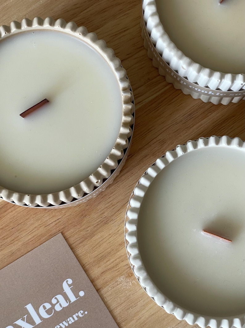 Puede incluir: Primer plano de tres velas de color crema en recipientes decorativos con bordes festoneados. Cada vela tiene una mecha de madera. Las velas est&aacute;n sobre una superficie de madera. Se ve una tarjeta con el texto "xleaf."