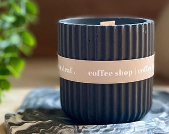 May include: A black, ribbed candle in a cylindrical concrete container with a wooden wick. The candle sits on a marbled gray and white tray. A tan band around the candle reads "coffee shop | coffee".