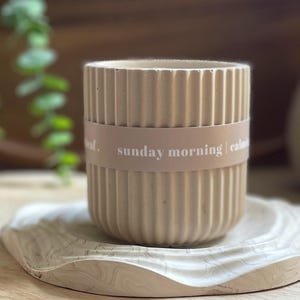 May include: A beige, ribbed ceramic container with a band that reads "sunday morning | calm". The container sits on a decorative, wavy-edged tray with a marbled pattern. The background includes greenery and a wooden surface.