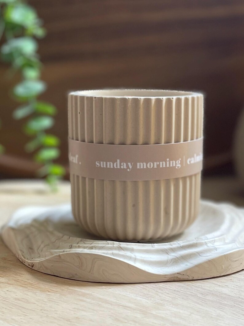 May include: A beige, ribbed ceramic container with a band that reads "sunday morning | calm". The container sits on a decorative, wavy-edged tray with a marbled pattern. The background includes greenery and a wooden surface.